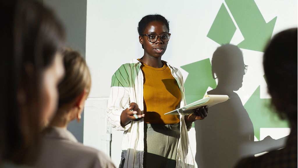 Junge afrikanische Frau hält einen Vortrag vor dem Hintergrund eines großen Recycling-Symbols. 