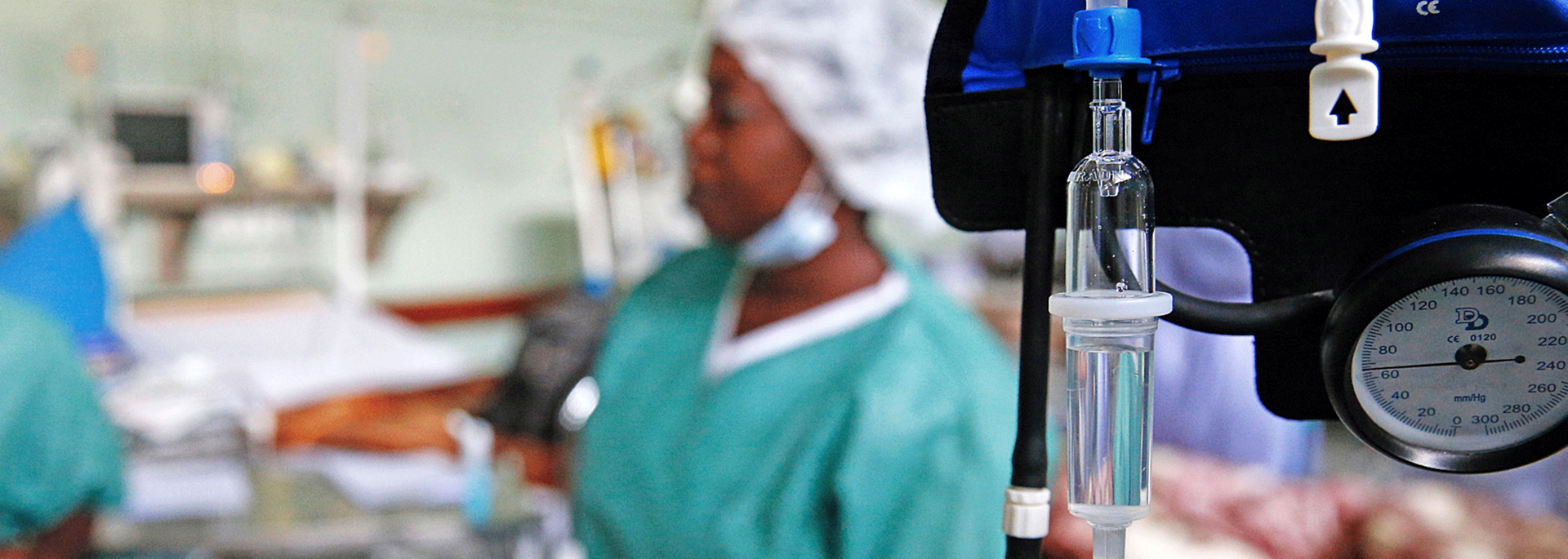 Innenaufnahme eines Krankenhauses in Afrika. Im Vordergrund ein Infusionsbeutel und ein Messgerät, im Hintergrund eine Krankenschwester.