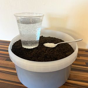 Produktfoto: Ein Teelöffel des Bodenhilfsstoffs Be-Grow mit einem Glas Wasser und Erde 